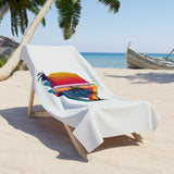 A white beach towel draped over a wooden lounge chair on a sandy beach, featuring a colorful tropical sunset and palm tree graphic.