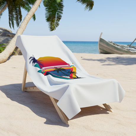 A white beach towel draped over a wooden lounge chair on a sandy beach, featuring a colorful tropical sunset and palm tree graphic.