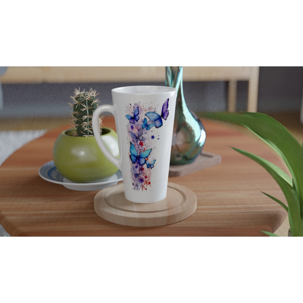 A tall white ceramic mug with a colorful watercolor butterfly and floral print, resting on a round wooden coaster on a wooden table. A small potted cactus and a decorative glass vase are visible in the softly blurred background.