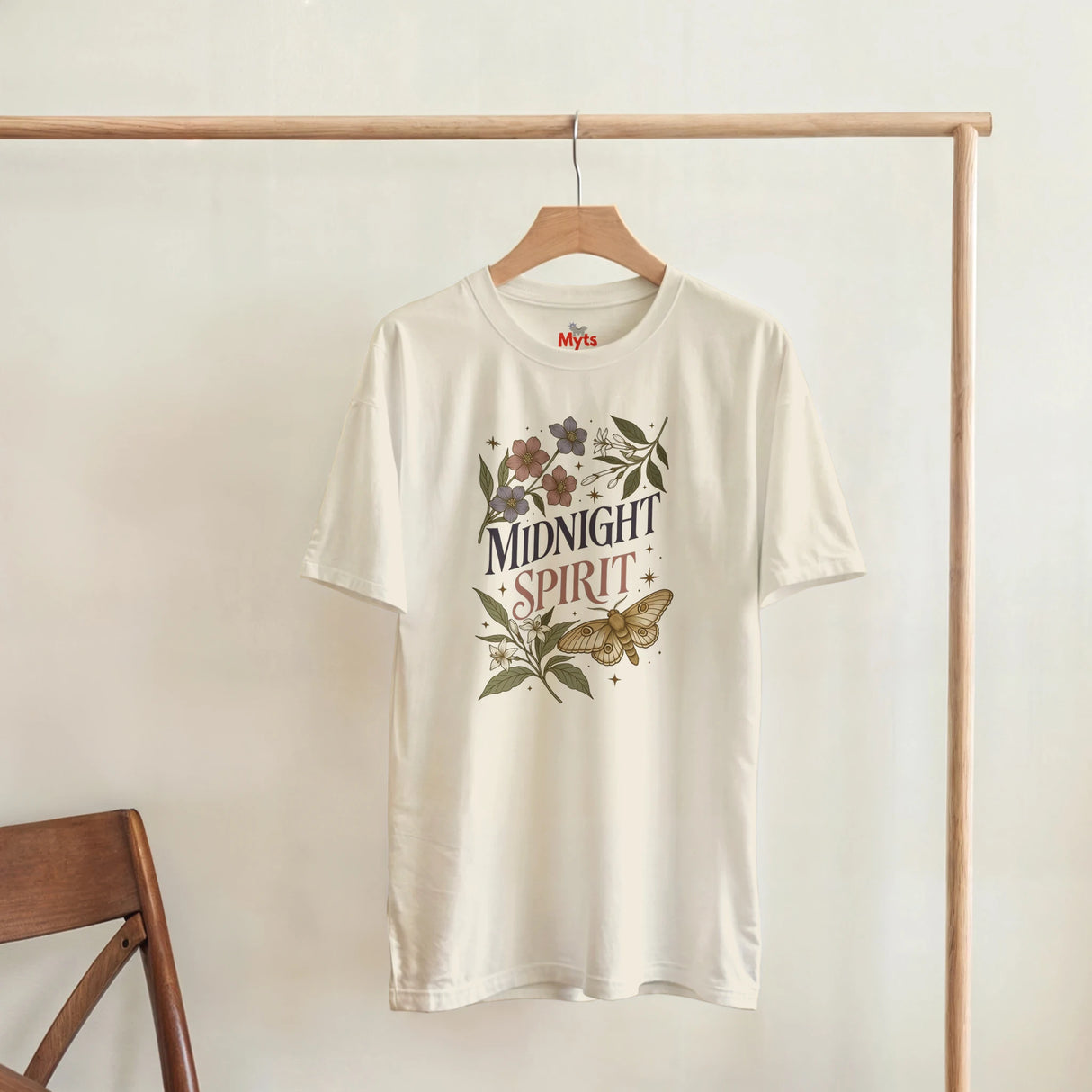 An off-white short-sleeve t-shirt hanging on a wooden hanger from a clothing rack. The shirt features a vintage-style graphic with the words 'MIDNIGHT SPIRIT' surrounded by flowers and a butterfly.