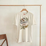 An off-white short-sleeve t-shirt hanging on a wooden hanger from a clothing rack. The shirt features a vintage-style graphic with the words 'MIDNIGHT SPIRIT' surrounded by flowers and a butterfly.