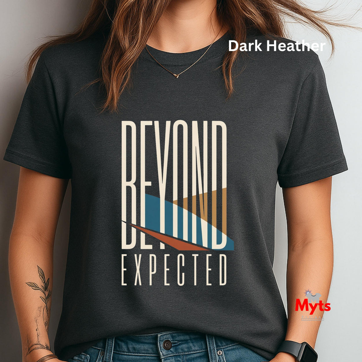 A woman wearing a dark heather charcoal t-shirt with a graphic print that reads 'BEYOND EXPECTED' in cream, gold, and blue. The text 'Dark Heather' is visible in the top right corner, and a watermark 'Myts' is visible in the bottom right corner.