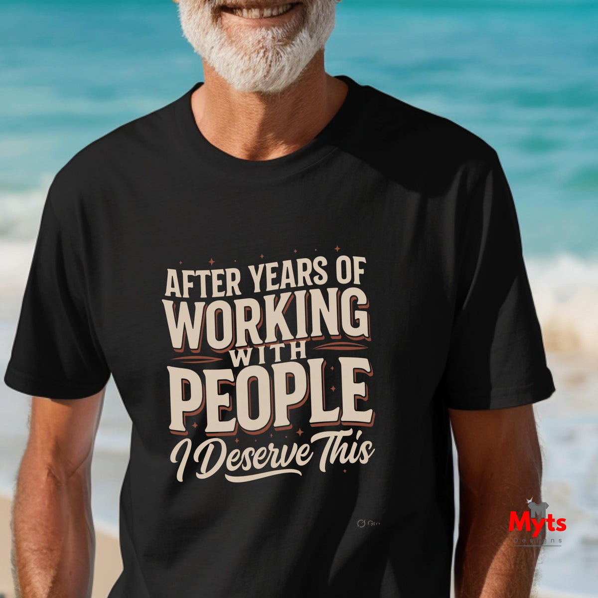 A man wearing a black graphic t-shirt with the text 'AFTER YEARS OF WORKING WITH PEOPLE I Deserve This' in beige and white lettering, set against a blurred beach background.