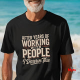 A man wearing a black graphic t-shirt with the text 'AFTER YEARS OF WORKING WITH PEOPLE I Deserve This' in beige and white lettering, set against a blurred beach background.