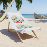 A white beach towel with colorful tropical coral and sea urchin embroidery, draped over a wooden lounge chair on a sandy beach with palm trees and the ocean in the background.