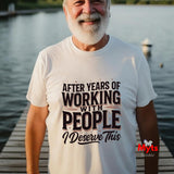 An older man with a white beard stands on a wooden dock by the water wearing a white graphic t-shirt with the text 'AFTER YEARS OF WORKING WITH PEOPLE I DESERVE THIS' in black and brown lettering. A red 'Myts' brand logo is visible in the bottom right corner.
