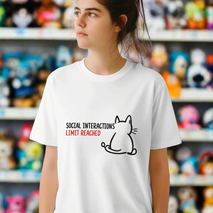“White T-shirt worn by a young woman ina store, with a minimal line art cat and text reading ‘Social Interactions Limit Reached’ in bold black and red font, humorous introvert design from MytsDesigns.com”
