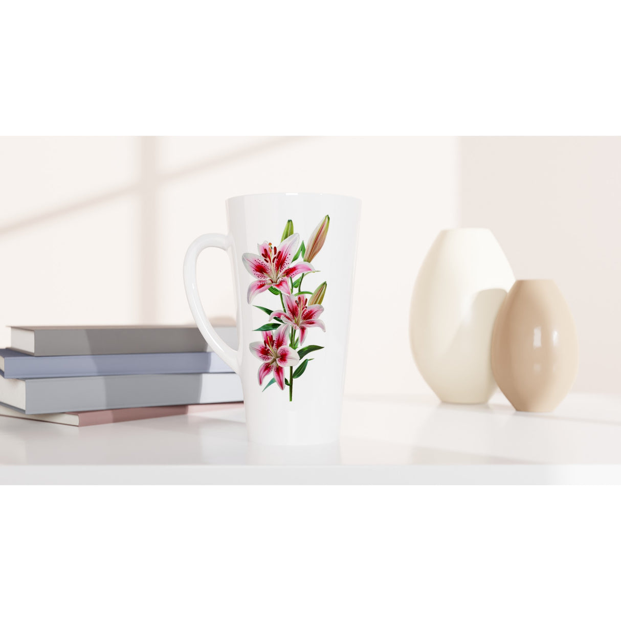 A tall white ceramic latte mug with a pink and green lily floral print, styled on a white surface next to stacked books and two ceramic vases.