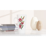 A tall white ceramic latte mug with a pink and green lily floral print, styled on a white surface next to stacked books and two ceramic vases.