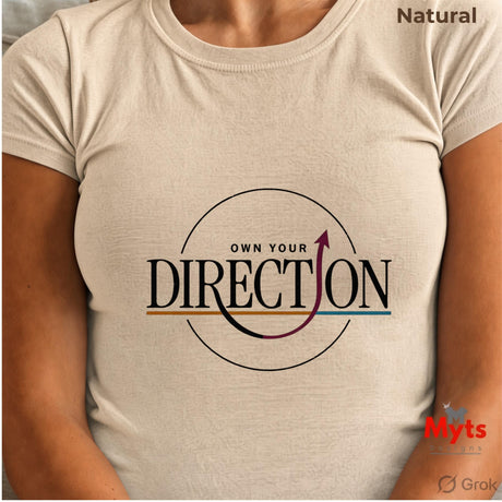 A person wearing a light natural beige short-sleeve t-shirt with a graphic print on the chest that reads 'OWN YOUR DIRECTION' inside a circular design with a curved arrow. The text 'Natural' is visible in the top right corner, and a watermark for 'Myts Designs' is in the bottom right.