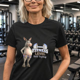 Woman in fitness centre wearing black Fitness Level Still Trying donkey graphic tee with exercise equipment behind her.