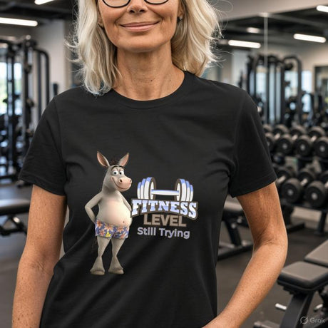 Woman in fitness centre wearing black Fitness Level Still Trying donkey graphic tee with exercise equipment behind her.