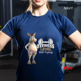 Woman in gym wearing Navy Fitness Level Still Trying graphic T-shirt with cartoon donkey and dumbbell artwork.