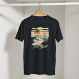 Black t-shirt with a beach graphic and 'The Shell' text on a hanger against a white wall.
