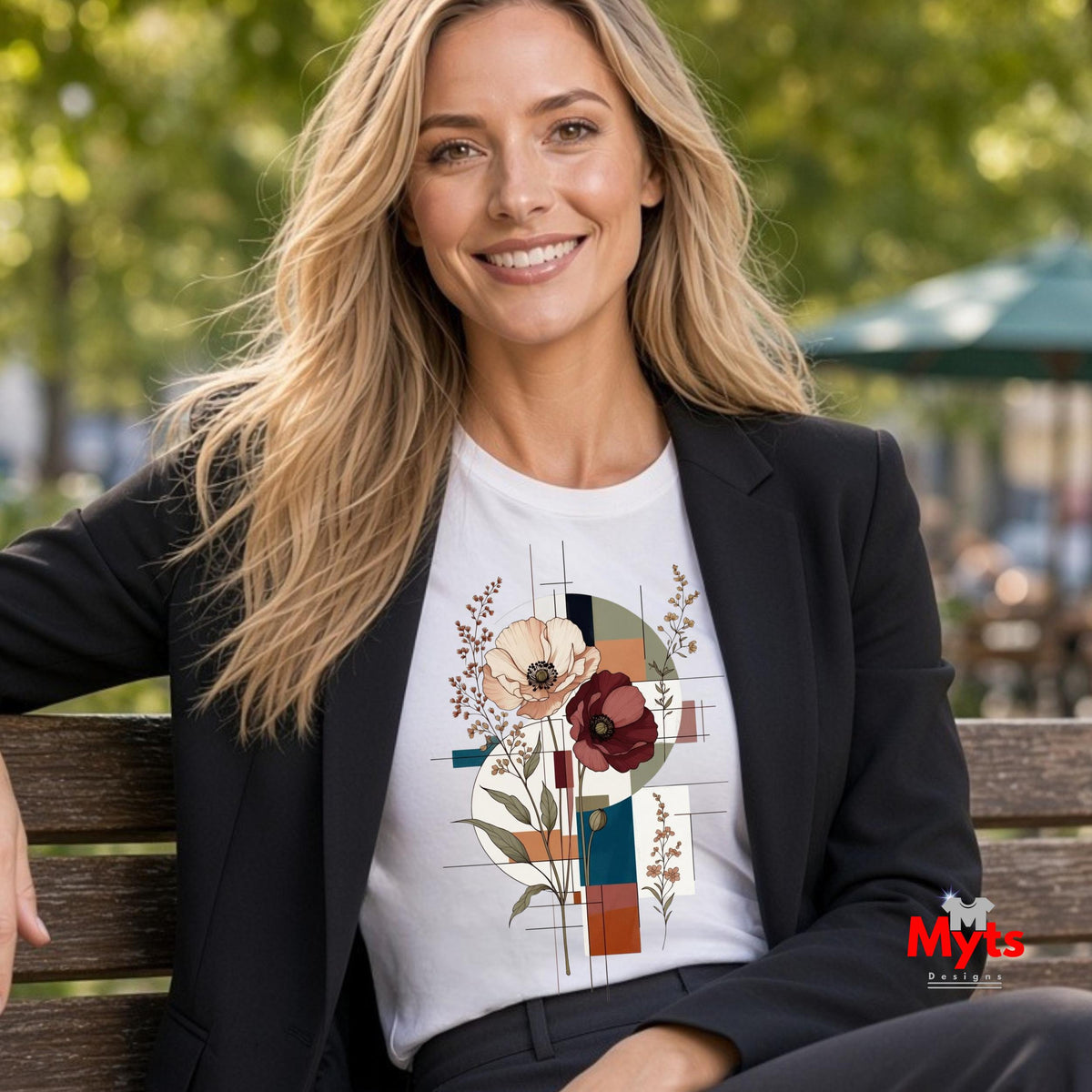 A woman sitting on a wooden bench outdoors wearing a white graphic t-shirt with a geometric floral print, styled with a black blazer. The background is a blurred park setting with green trees.