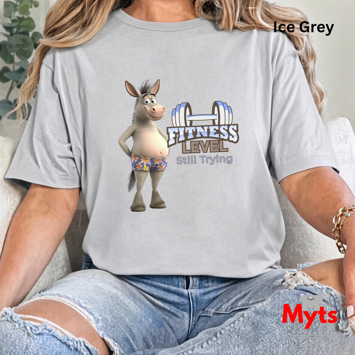 Woman seated in casual jeans wearing Ice Grey Fitness Level Still Trying humorous donkey graphic T-shirt.