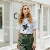 Woman wearing a white t-shirt with a graphic design and green pants in an indoor setting.