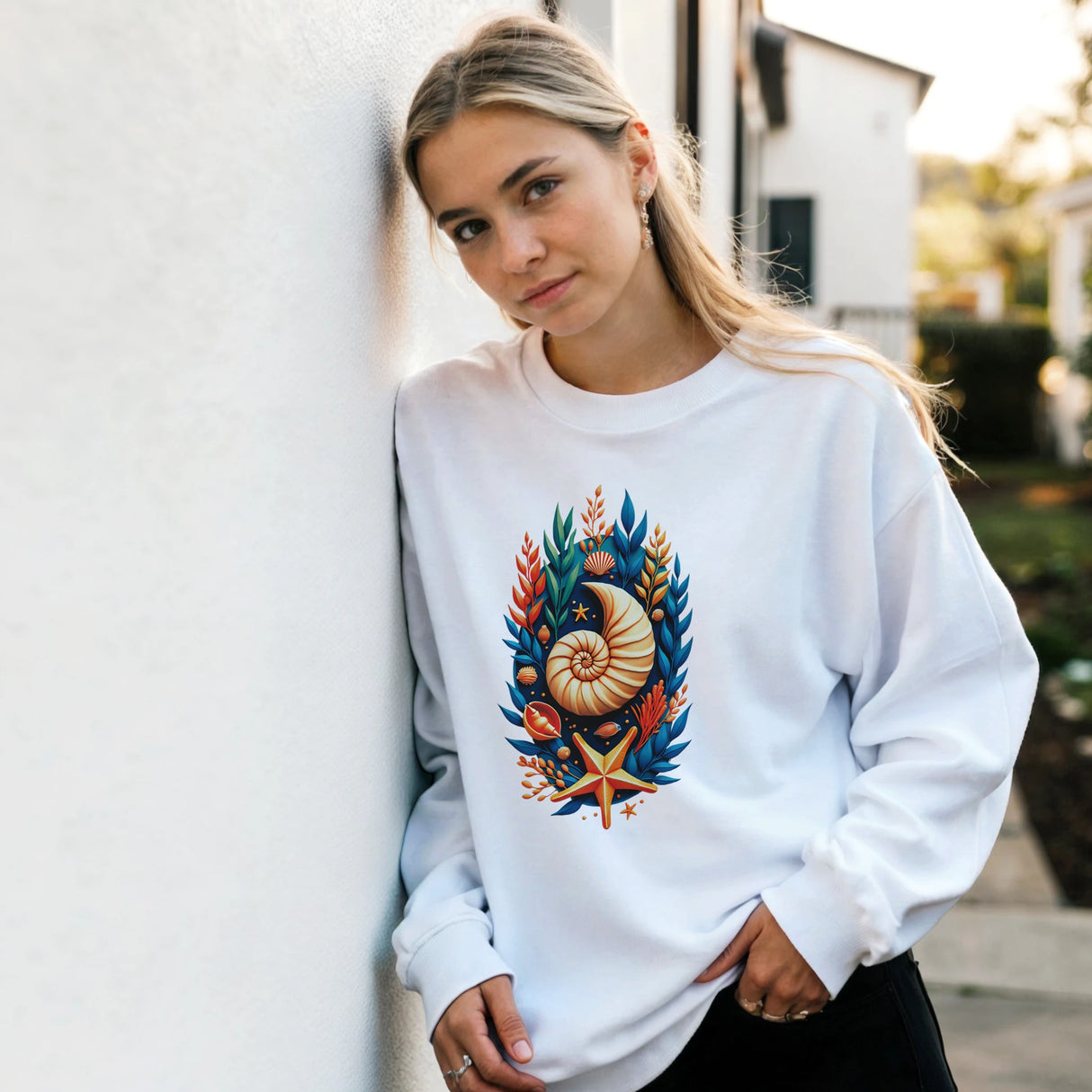 A woman wearing a white crewneck sweatshirt with a colorful seashell and starfish graphic, leaning against a white stucco wall outdoors.