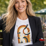 A smiling blonde woman wearing a black blazer over a white graphic T-shirt, seated on a wooden bench outdoors with blurred greenery in the background. The shirt features a large abstract orange, tan, and dark blue graphic with the word 'UNBOUND' at the bottom.