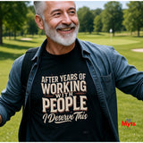 A smiling older man with gray hair and a beard stands outdoors on a grassy golf course wearing a black graphic t-shirt under an open blue denim shirt. The t-shirt features the text 'AFTER YEARS OF WORKING WITH PEOPLE I Deserve This' in beige and white lettering.