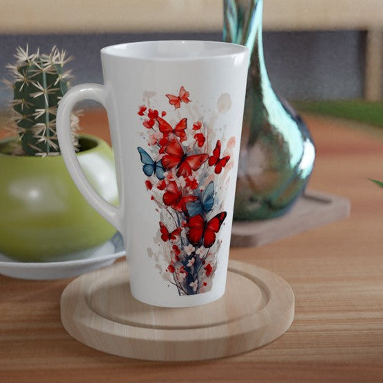 A tall white ceramic mug with a red and blue butterfly and floral print, resting on a round wooden coaster on a wooden table. A green potted cactus and a blue glass vase are visible in the background.