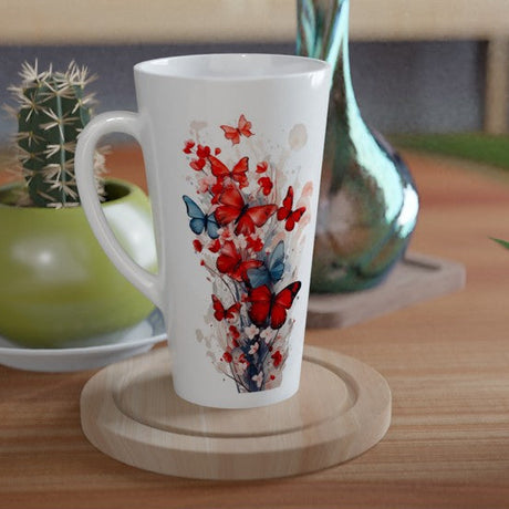 A tall white ceramic mug with a red and blue butterfly and floral print, resting on a round wooden coaster on a wooden table. A green potted cactus and a blue glass vase are visible in the background.