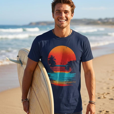 Man holding a surfboard on a beach wearing a navy t-shirt with a colorful sunset design.