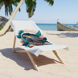 A white beach towel featuring a colorful tropical fish and coral graphic print, draped over a wooden lounge chair on a sandy beach with palm trees and the ocean in the background.