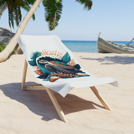 A white beach towel featuring a colorful tropical fish and coral graphic print, draped over a wooden lounge chair on a sandy beach with palm trees and the ocean in the background.