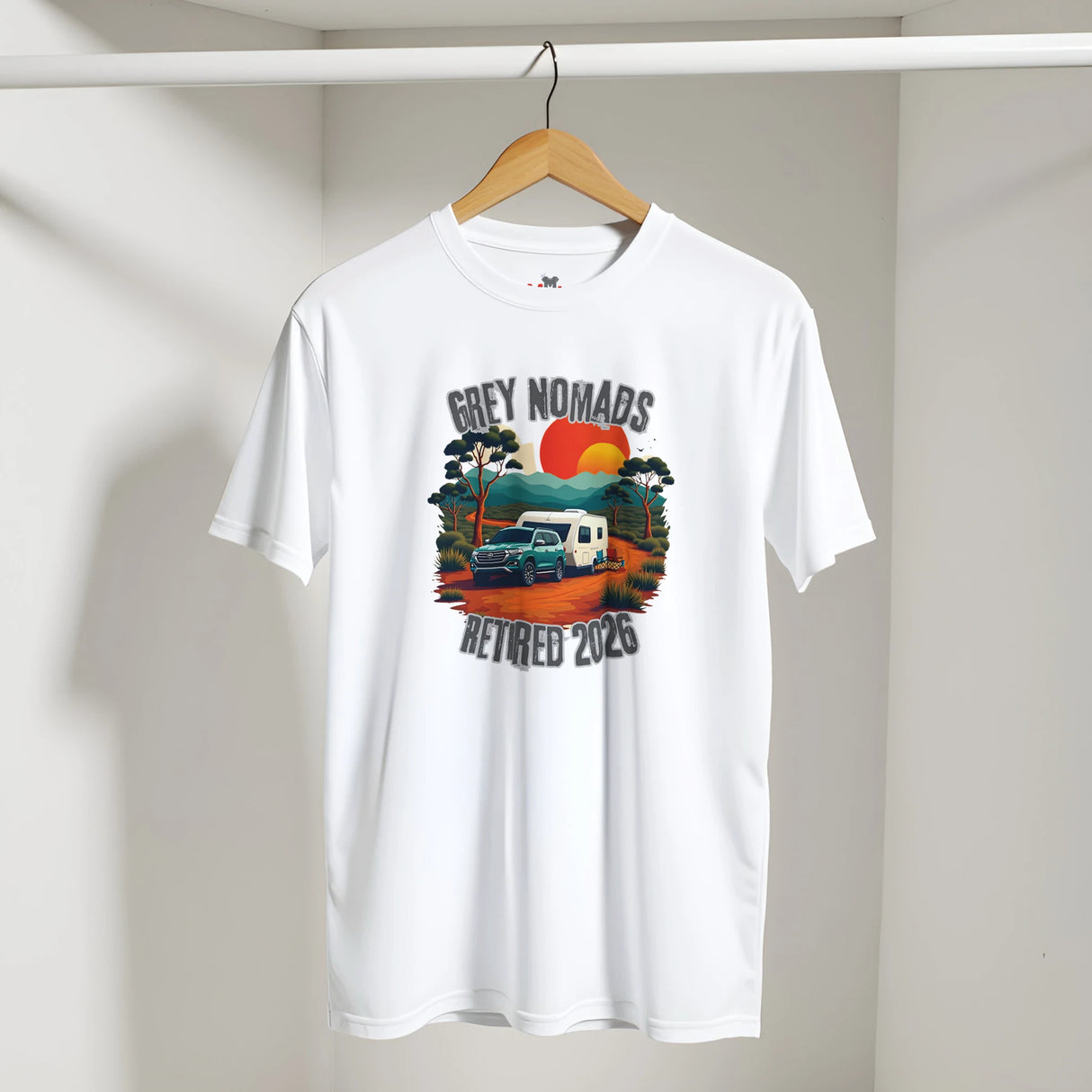 A white short-sleeve t-shirt hanging on a wooden hanger against a light gray wall. The shirt features a colorful retro-style graphic in the center with the text 'GREY NOMADS RETIRED 2023' surrounding an illustration of a blue truck, a white camper van, trees, and a red and orange sun.