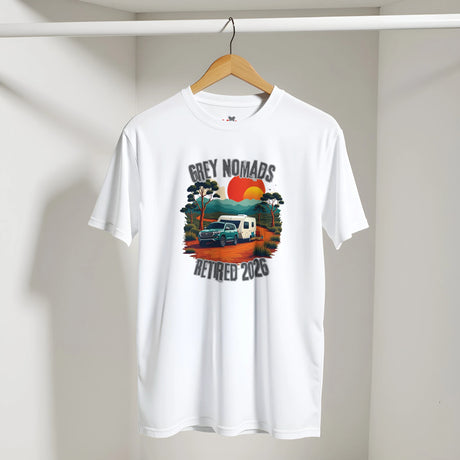 A white short-sleeve t-shirt hanging on a wooden hanger against a light gray wall. The shirt features a colorful retro-style graphic in the center with the text 'GREY NOMADS RETIRED 2023' surrounding an illustration of a blue truck, a white camper van, trees, and a red and orange sun.