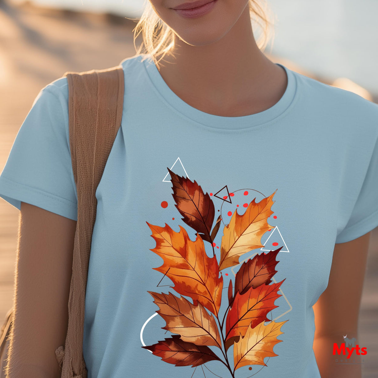 A woman wearing a light blue crewneck t-shirt with a graphic print of autumn maple leaves in orange, red, and brown, styled with geometric white line art and red dots. She has a tan shoulder bag and is photographed outdoors with a blurred sandy background.