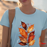 A woman wearing a light blue crewneck t-shirt with a graphic print of autumn maple leaves in orange, red, and brown, styled with geometric white line art and red dots. She has a tan shoulder bag and is photographed outdoors with a blurred sandy background.