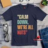 A flat lay view of a navy retro styled t-shirt on wooden boards, featuring distressed lettering "Calm Down, We're All Nuts"