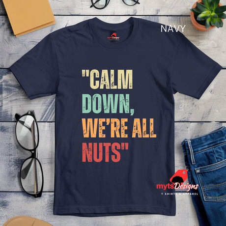 A flat lay view of a navy retro styled t-shirt on wooden boards, featuring distressed lettering "Calm Down, We're All Nuts"