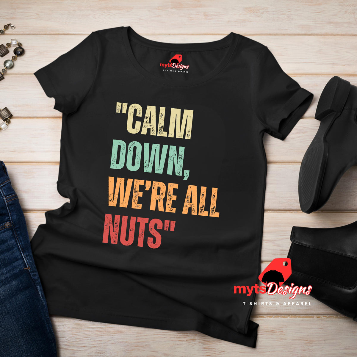 A flat lay view of a black retro styled t-shirt on wooden boards, featuring distressed lettering "Calm Down, We're All Nuts"