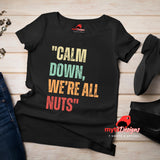 A flat lay view of a black retro styled t-shirt on wooden boards, featuring distressed lettering "Calm Down, We're All Nuts"