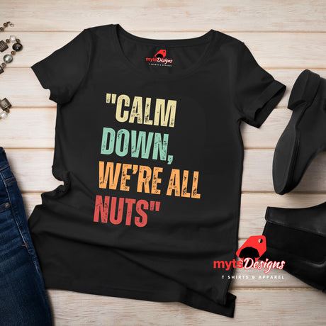 A flat lay view of a black retro styled t-shirt on wooden boards, featuring distressed lettering "Calm Down, We're All Nuts"