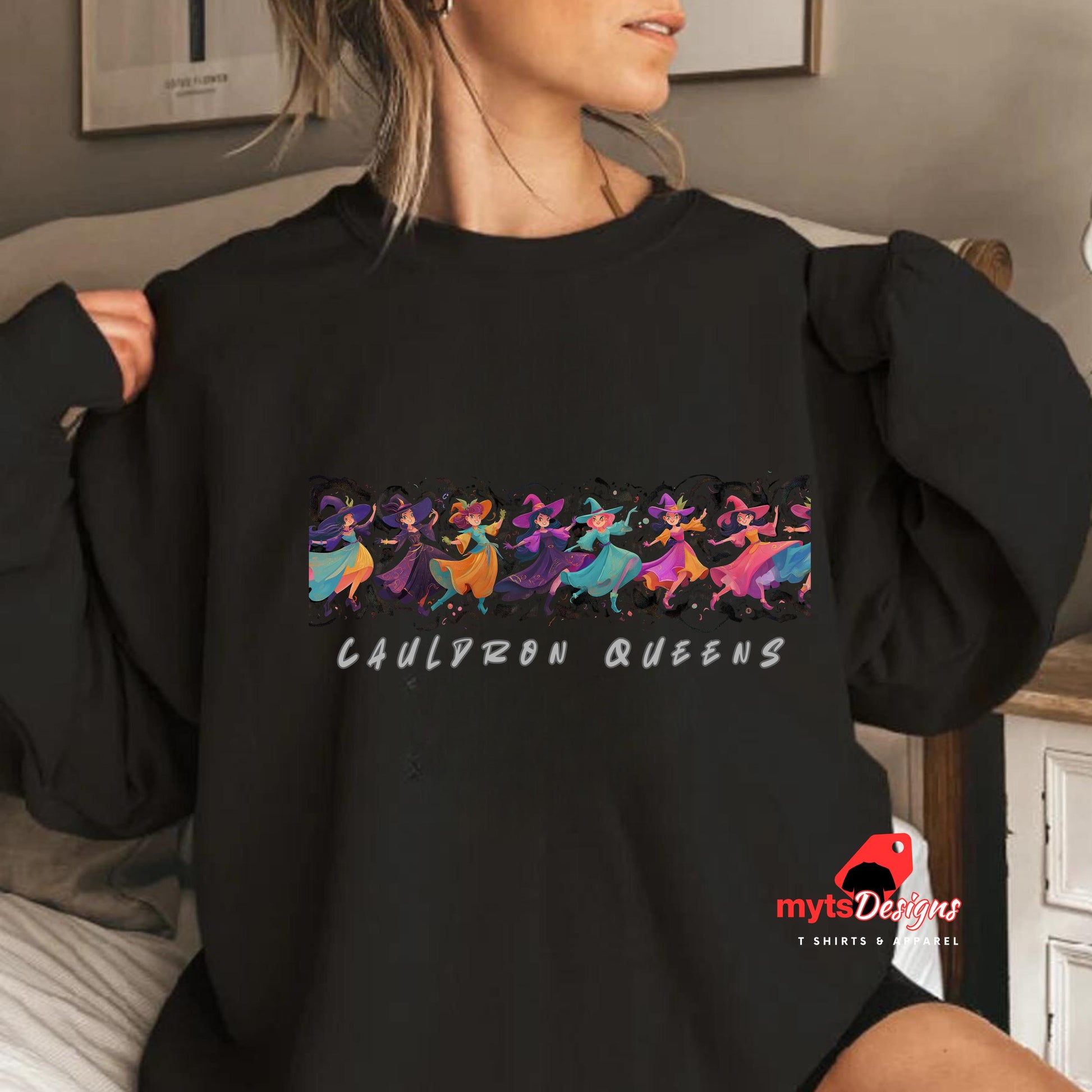 Person wearing a black sweatshirt with 'Cauldron Queens' design and colorful characters.