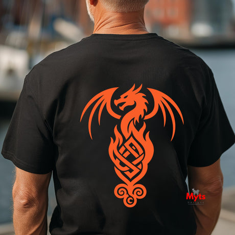A man viewed from behind wearing a black t-shirt with a large orange Celtic dragon graphic printed on the back. The background is a blurred outdoor setting with water and boats.