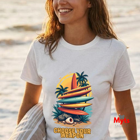 Person wearing a white t-shirt with a colorful graphic design of surfboards and palm trees, standing on a beach.