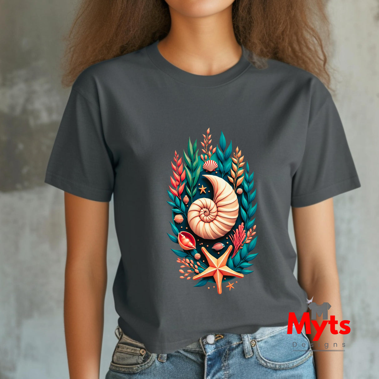 A woman wearing a dark gray t-shirt with a colorful graphic of a nautilus shell, starfish, and seashells. The background is a textured gray wall.