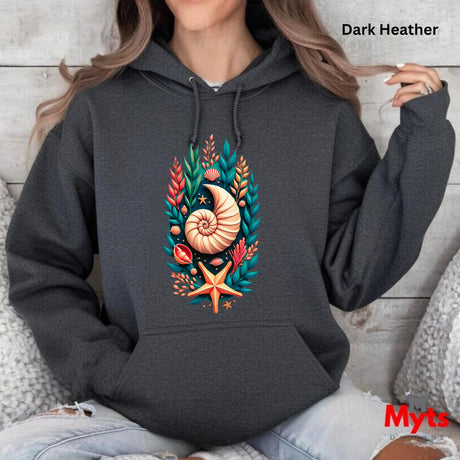 A woman wearing a dark heather charcoal hoodie with a colorful seashell and coral graphic on the front, sitting against a light wood wall. Text overlays read 'Dark Heather' and 'Myts'.