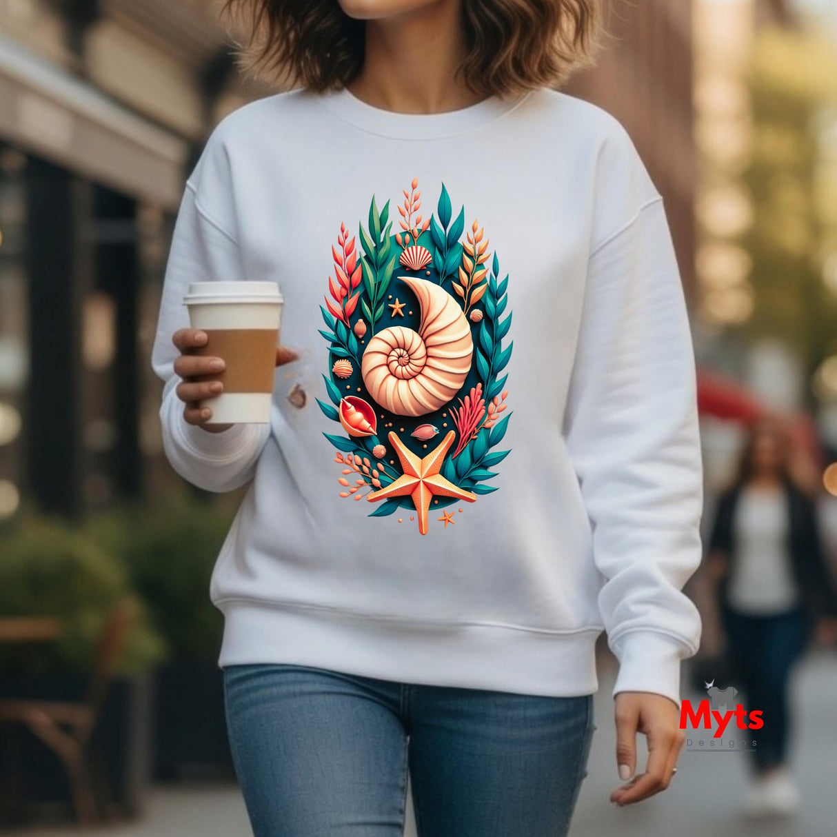 A woman wearing a white crewneck sweatshirt with a colorful graphic print of a nautilus shell, starfish, and tropical leaves, holding a coffee cup in an outdoor urban setting.