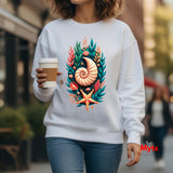 A woman wearing a white crewneck sweatshirt with a colorful graphic print of a nautilus shell, starfish, and tropical leaves, holding a coffee cup in an outdoor urban setting.