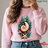 A woman wearing a light pink crewneck sweatshirt with a colorful nautical graphic on the front, featuring a spiral shell, starfish, coral, and green leaves. She is holding a phone to take a mirror selfie, with blue jeans visible at the bottom.