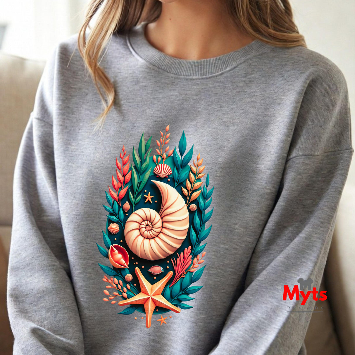 A person wearing a heather gray crewneck sweatshirt with a colorful graphic of a nautilus shell, starfish, and seashells on the front. The photo is cropped from the neck down, showing the sweatshirt and the model's hair against a soft indoor background.