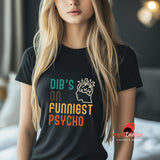Front view of a young woman wearing a casual tee featuring retro script that reads “Dibs on Funniest Psycho” in distressed vintage style, printed on ultra-soft black Gildan 64000 cotton
