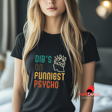 Front view of a young woman wearing a casual tee featuring retro script that reads “Dibs on Funniest Psycho” in distressed vintage style, printed on ultra-soft black Gildan 64000 cotton