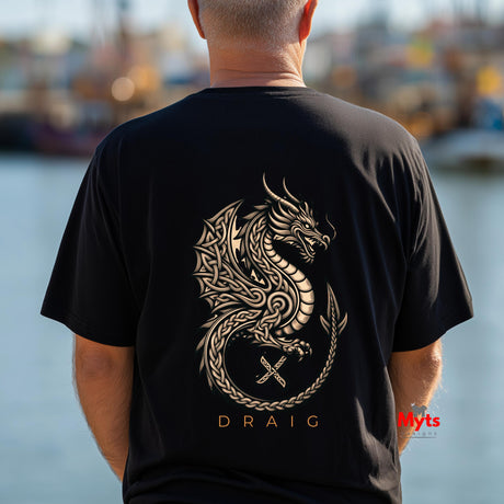 A man viewed from behind wearing a black t-shirt with a large gold Celtic-style dragon graphic and the name 'DRAIG' printed below it, set against a blurred waterfront background.